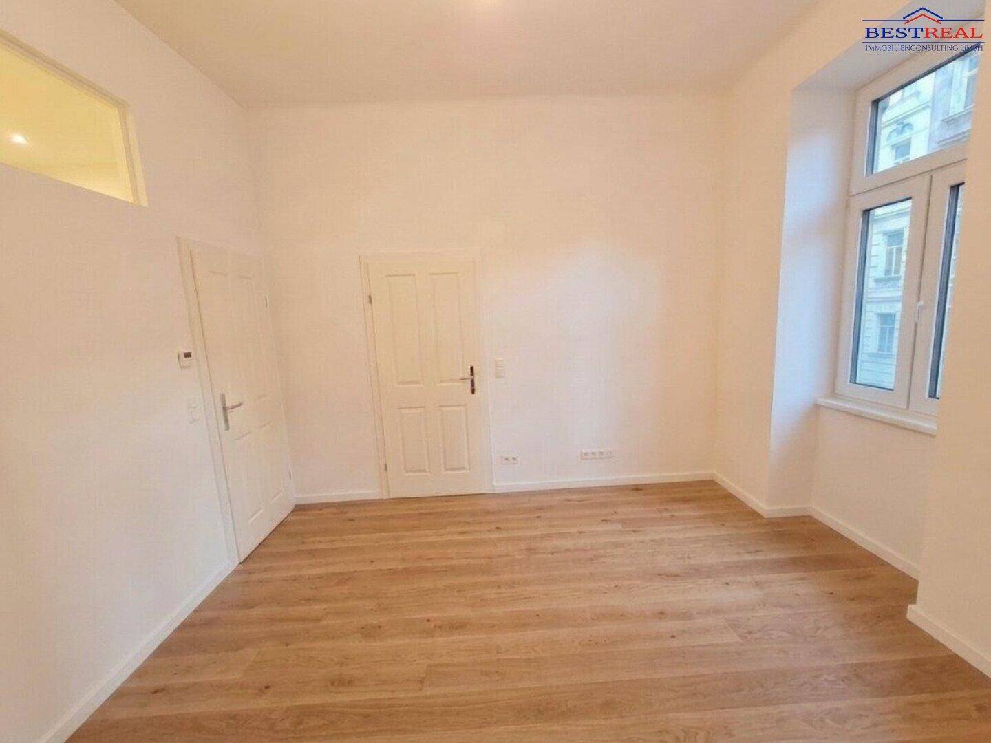Geräumiges Zimmer mit Holzparkettboden und einem großen Fenster, frisch renoviert und hell.
