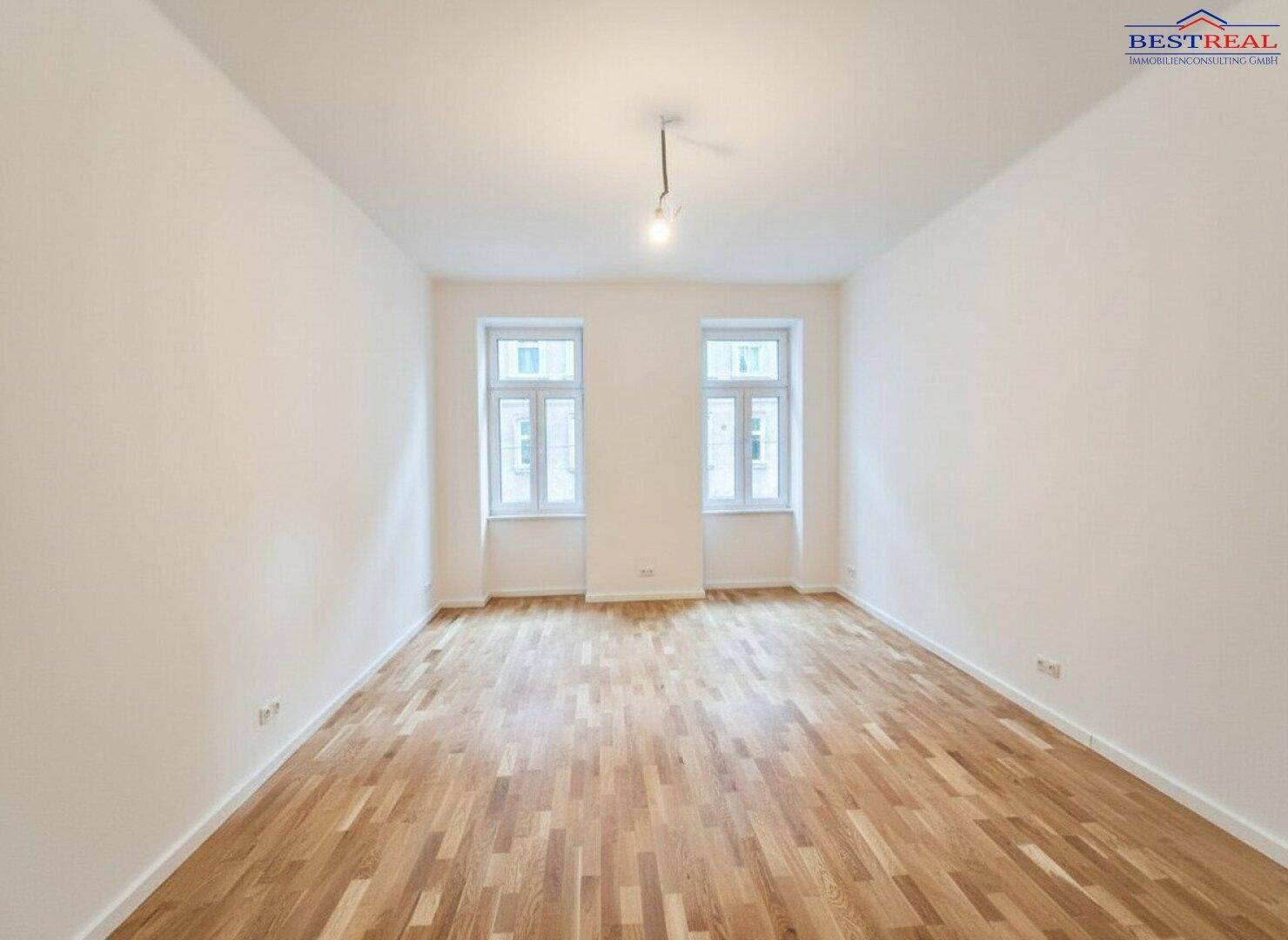 Geräumiger Wohnraum mit hellem Parkettboden und zwei großen Fenstern, lichtdurchflutet.