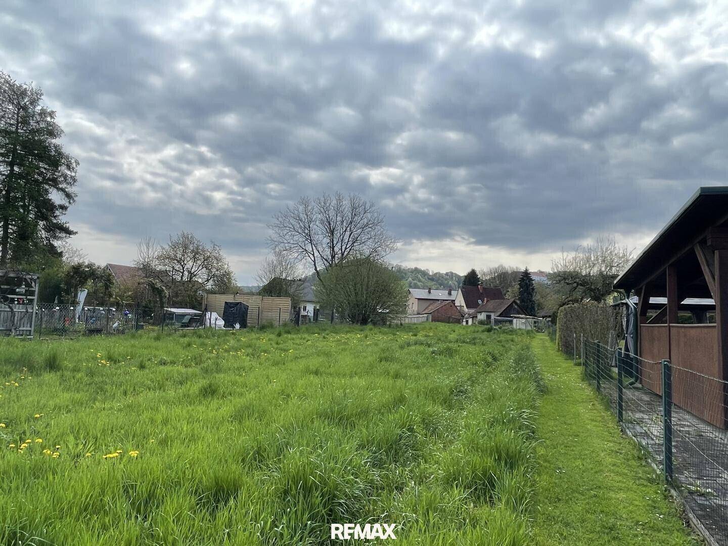 Langes, grünes Grundstück mit hohem Gras und Blick auf die Nachbarhäuser unter einem bewölkten Himmel.