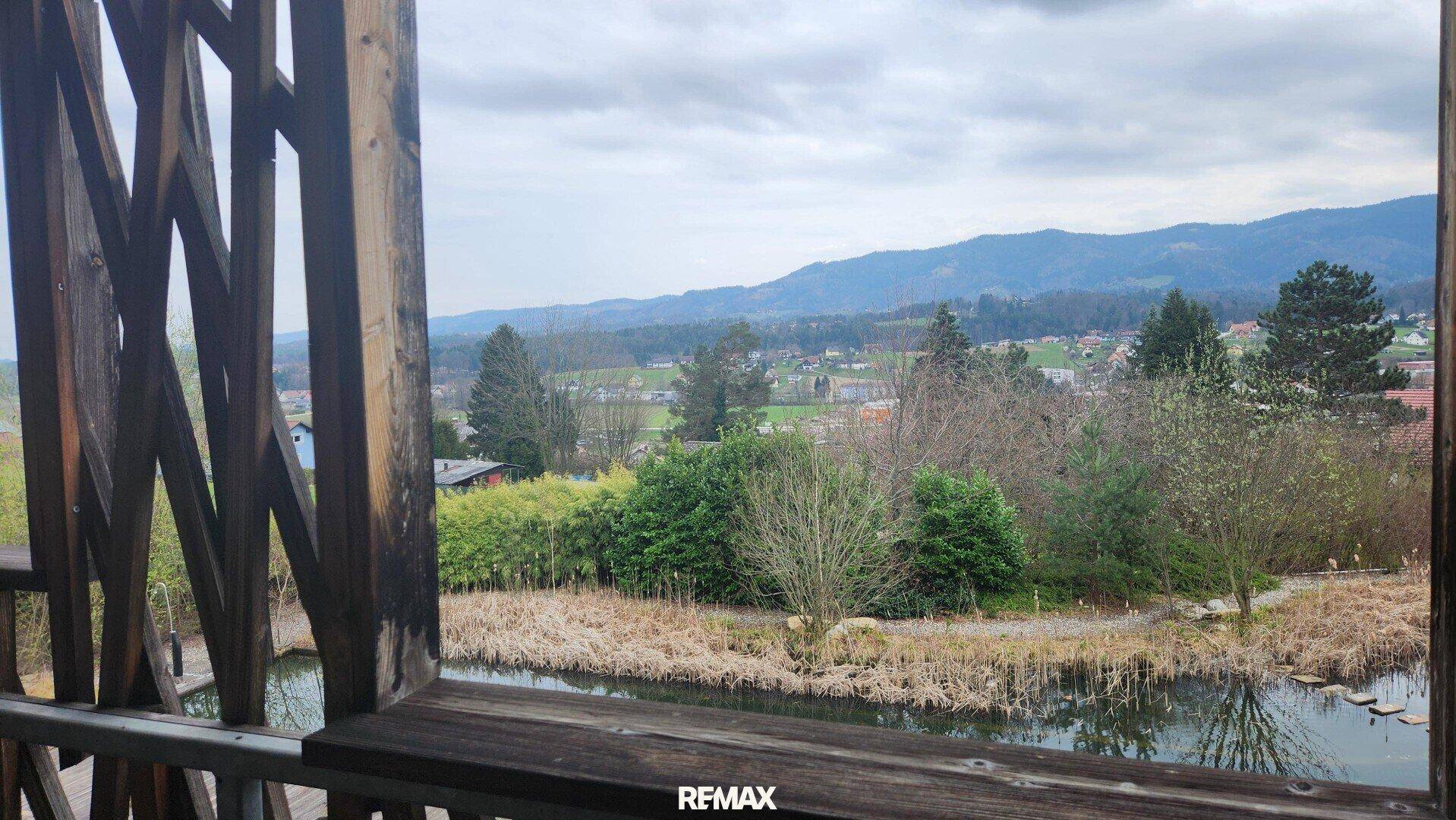 Blick von einem Holzbalkon auf einen Teich, grüne Landschaft und ein Dorf mit Bergen im Hintergrund.