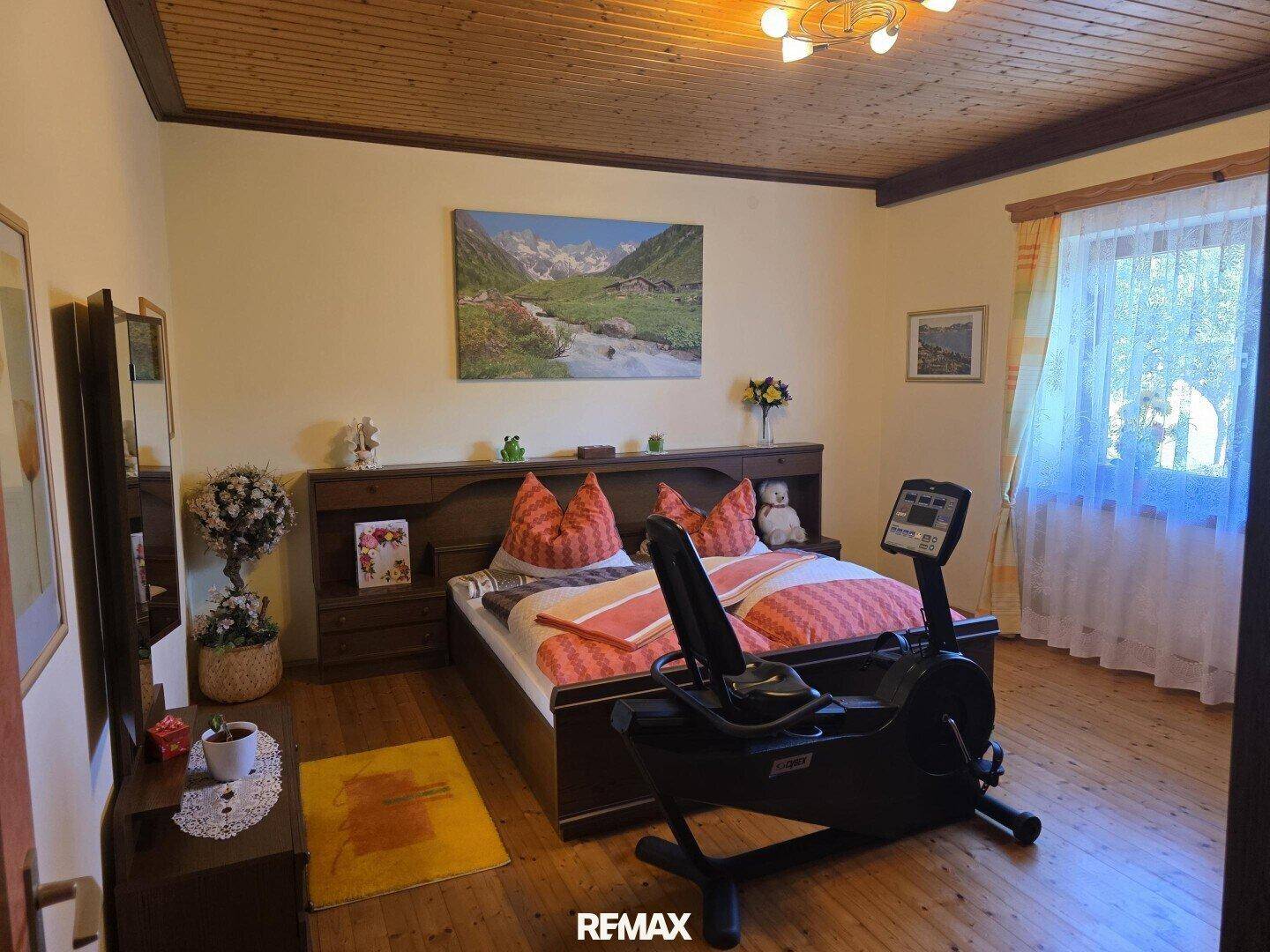 Gemütliches Schlafzimmer mit Holzboden, Holzdecke und einem Heimtrainer am Fenster.