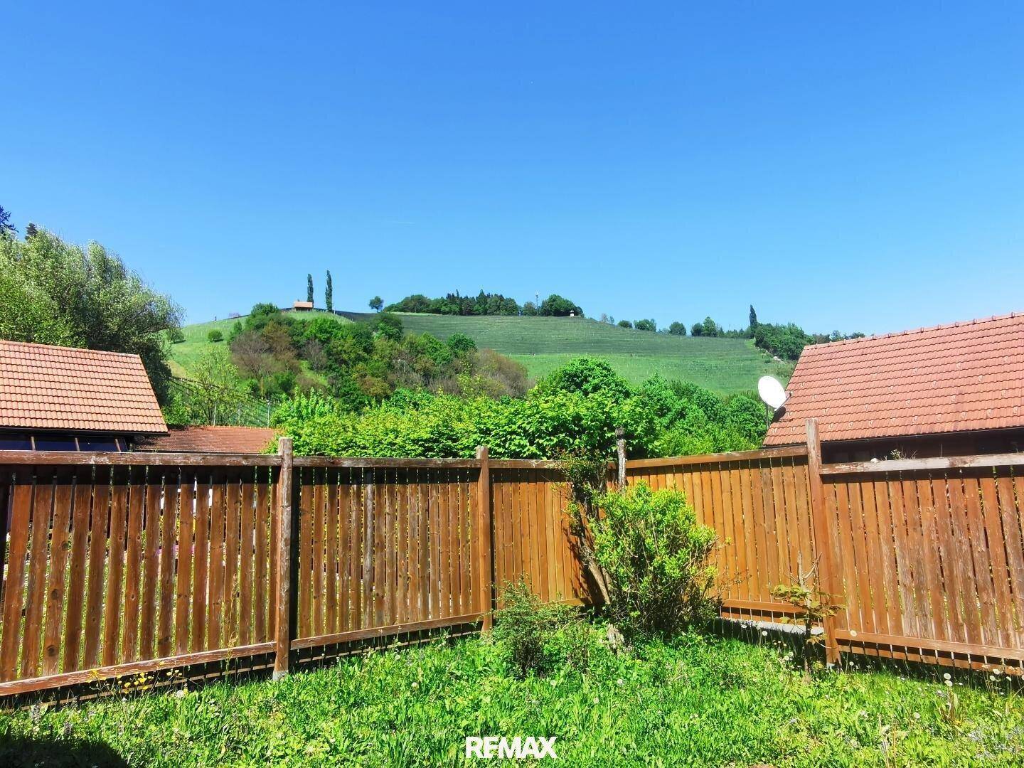 Eingezäunter Gartenbereich mit Rasenfläche und Blick auf eine grüne Hügellandschaft unter blauem Himmel.