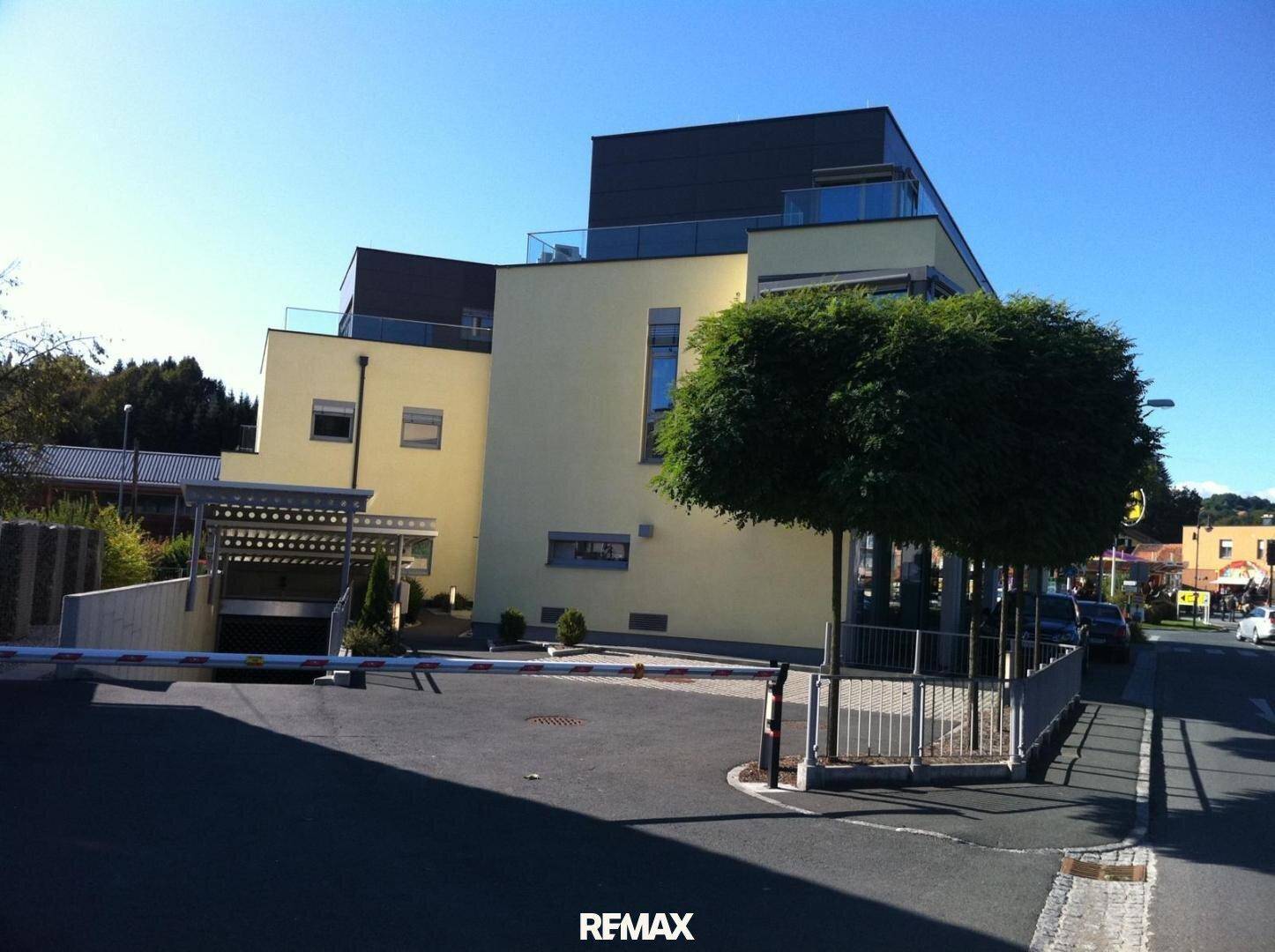 Seitenansicht des modernen Gebäudes mit gelber Fassade und der Einfahrt zur Tiefgarage unter blauem Himmel.