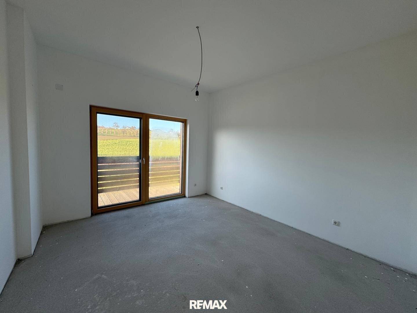 Unmöblierter Raum im Rohbau mit Zugang zu einem Balkon und Blick ins Grüne.