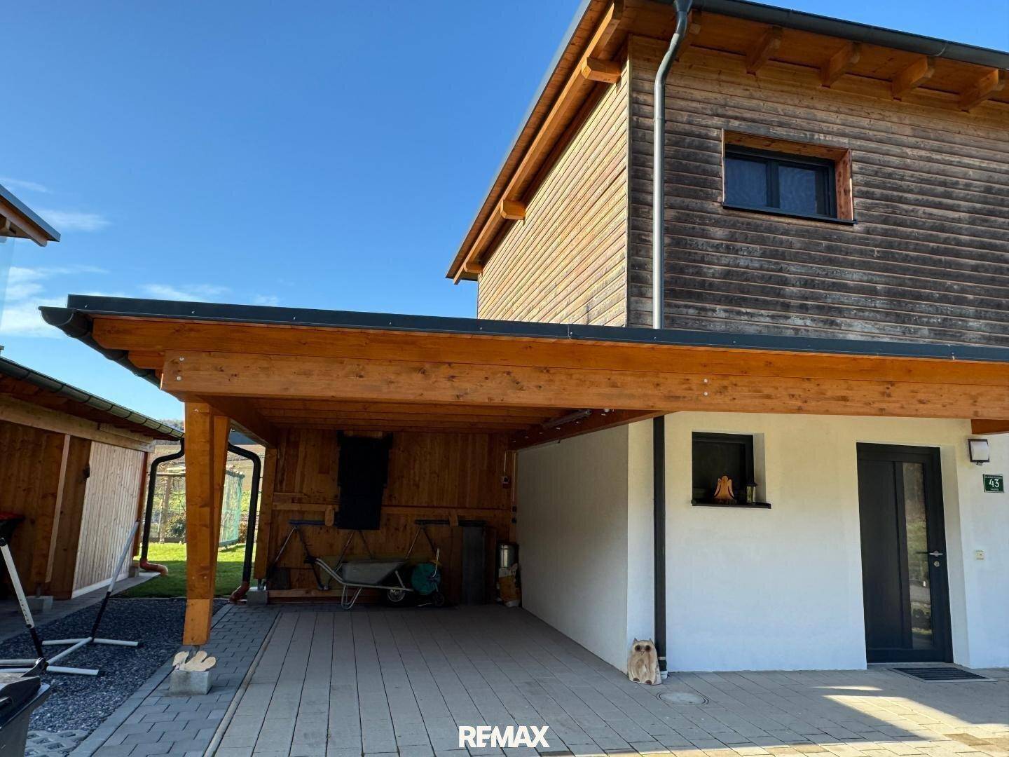 Holzcarport neben dem Haus mit gepflastertem Stellplatz und angrenzendem Geräteschuppen.