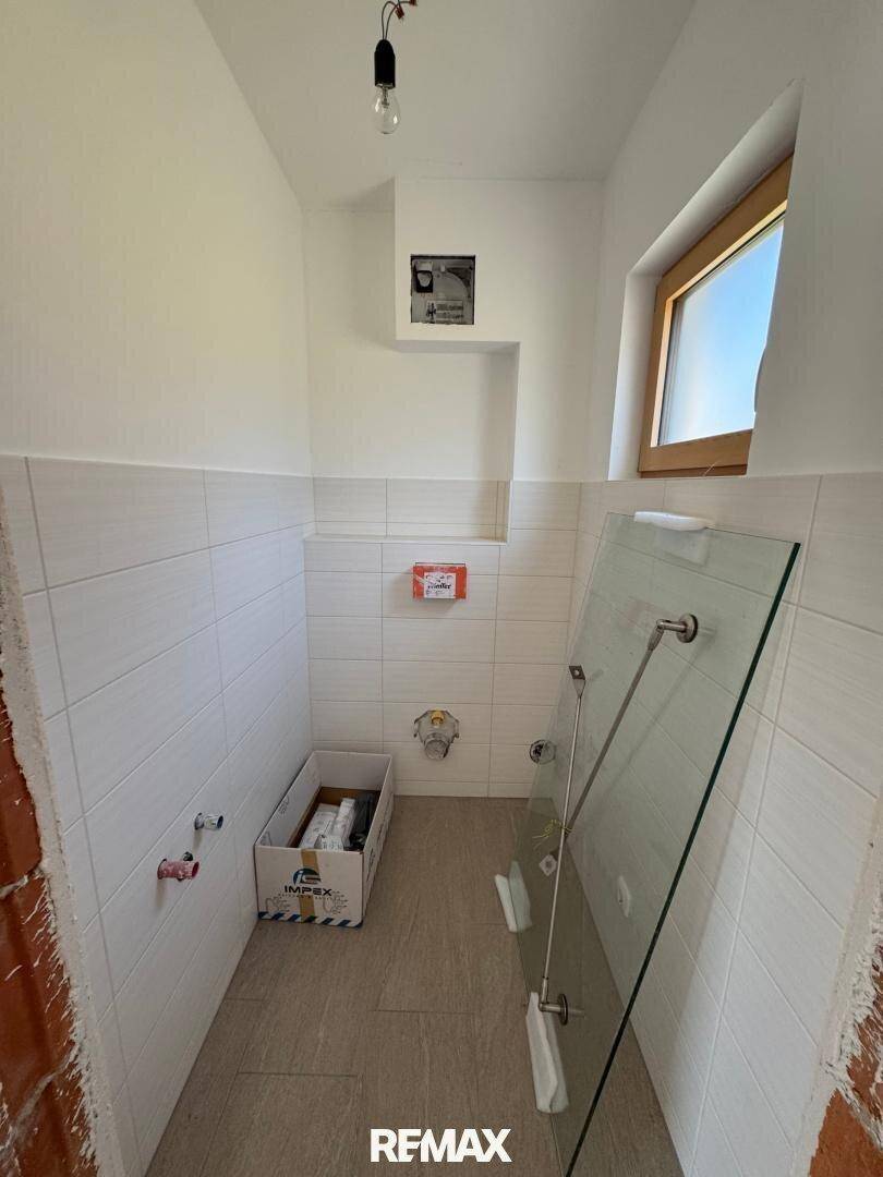 Unfertiges Gäste-WC mit Fliesen, Fenster und vorbereiteten Anschlüssen für Sanitäranlagen.