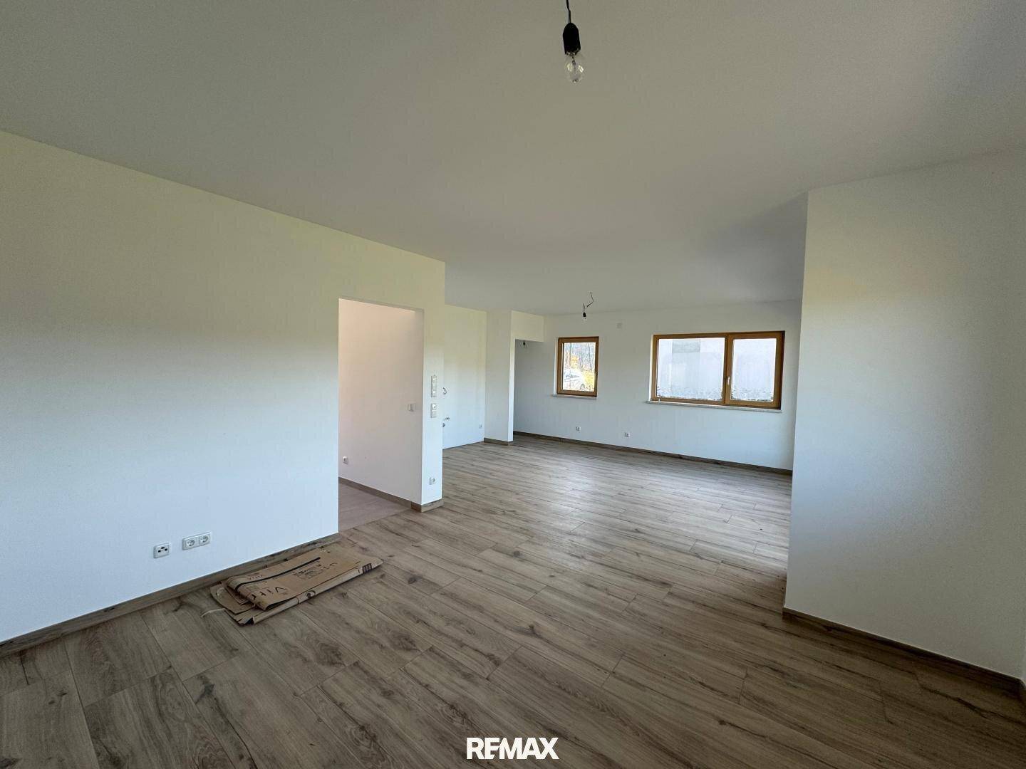 Geräumiger, unmöblierter Wohnbereich im Rohbau mit großen Fenstern und Holzboden.