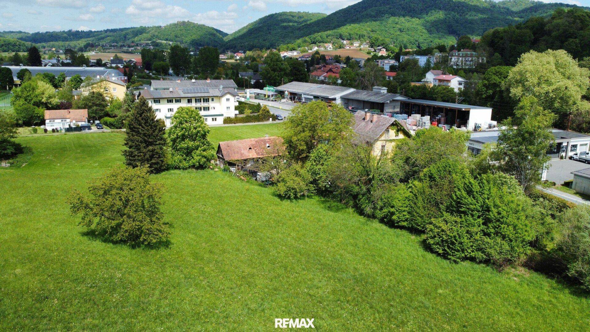 Luftaufnahme eines weitläufigen grünen Grundstücks mit Häusern, Gewerbe und Hügeln im Hintergrund.