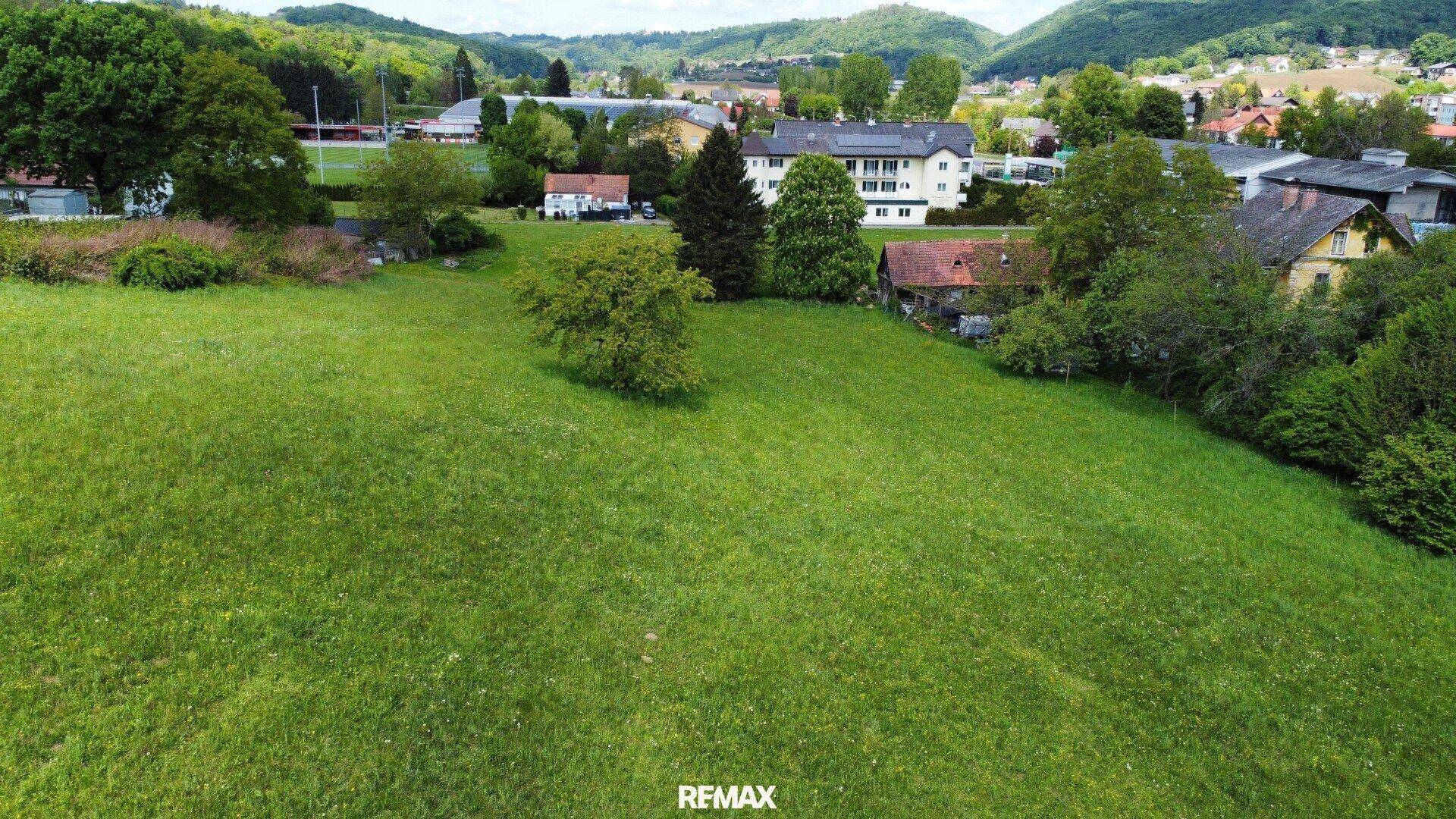 Luftaufnahme eines weitläufigen grünen Grundstücks mit Häusern und bewaldeten Hügeln im Hintergrund.