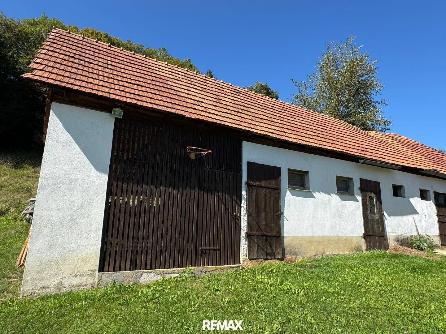 Ländliches Gebäude mit weißer Fassade, rotem Ziegeldach und großen Holztüren in grüner Umgebung.