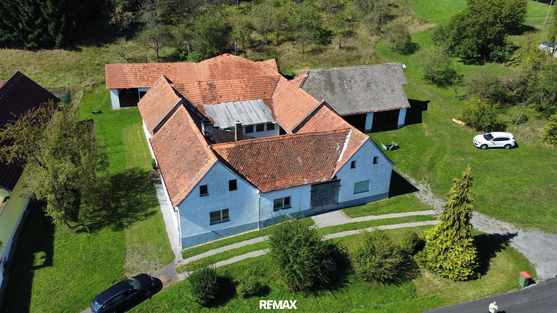 Vogelperspektive eines Bauernhofs mit mehreren Gebäuden und umgebenden Feldern und Wäldern.