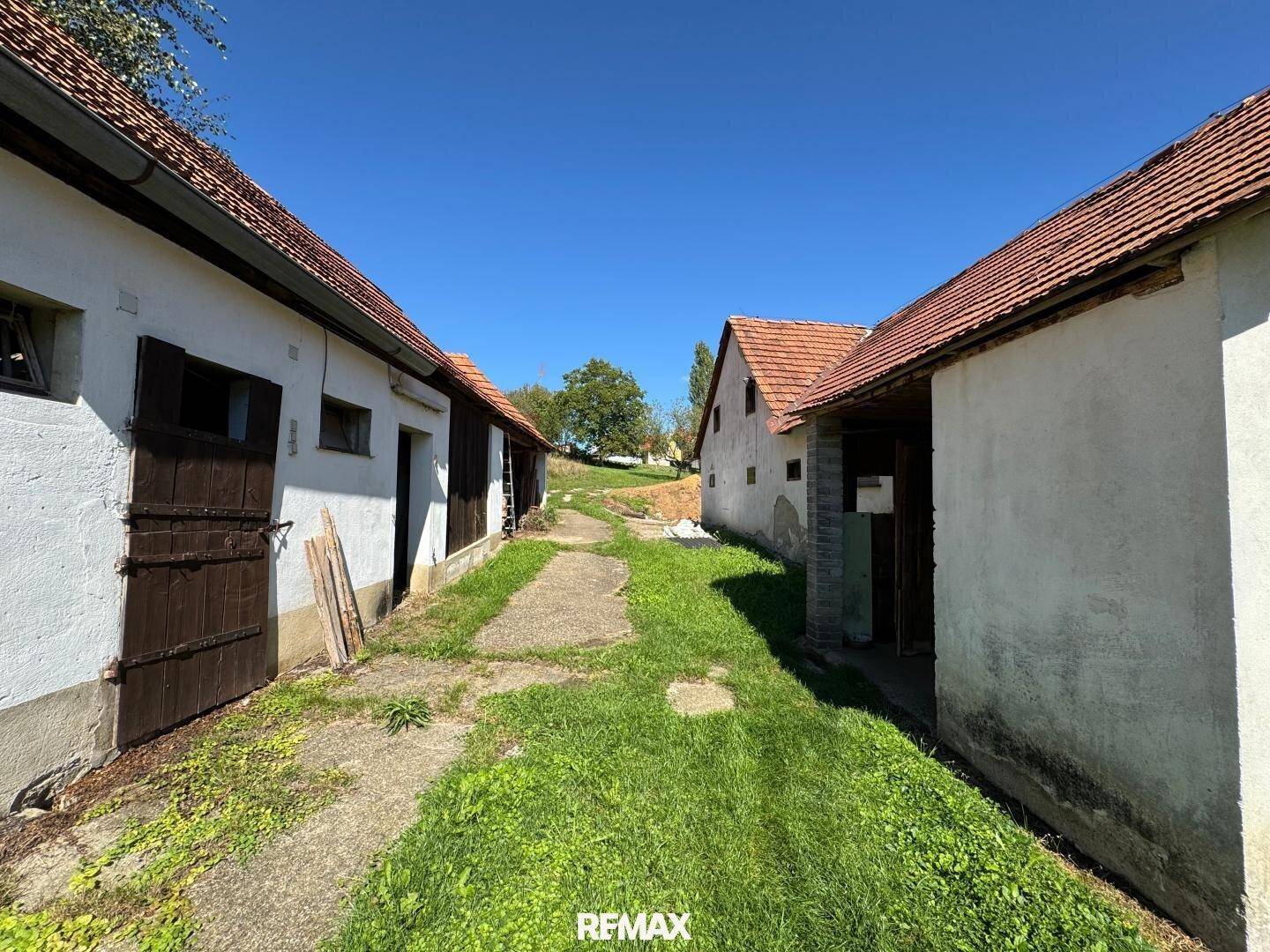Zwei ländliche Gebäude mit roten Ziegeldächern und einem grasbewachsenen Weg unter blauem Himmel.