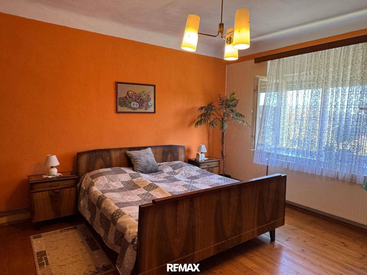 Schlafzimmer mit orangefarbenen Wänden, Holzbett und einem großen Fenster mit Vorhängen.