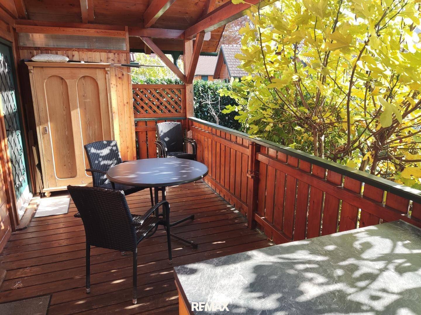 Überdachter Holzbalkon mit Esstisch und Stühlen, umgeben von herbstlicher Vegetation und Sonnenschein.