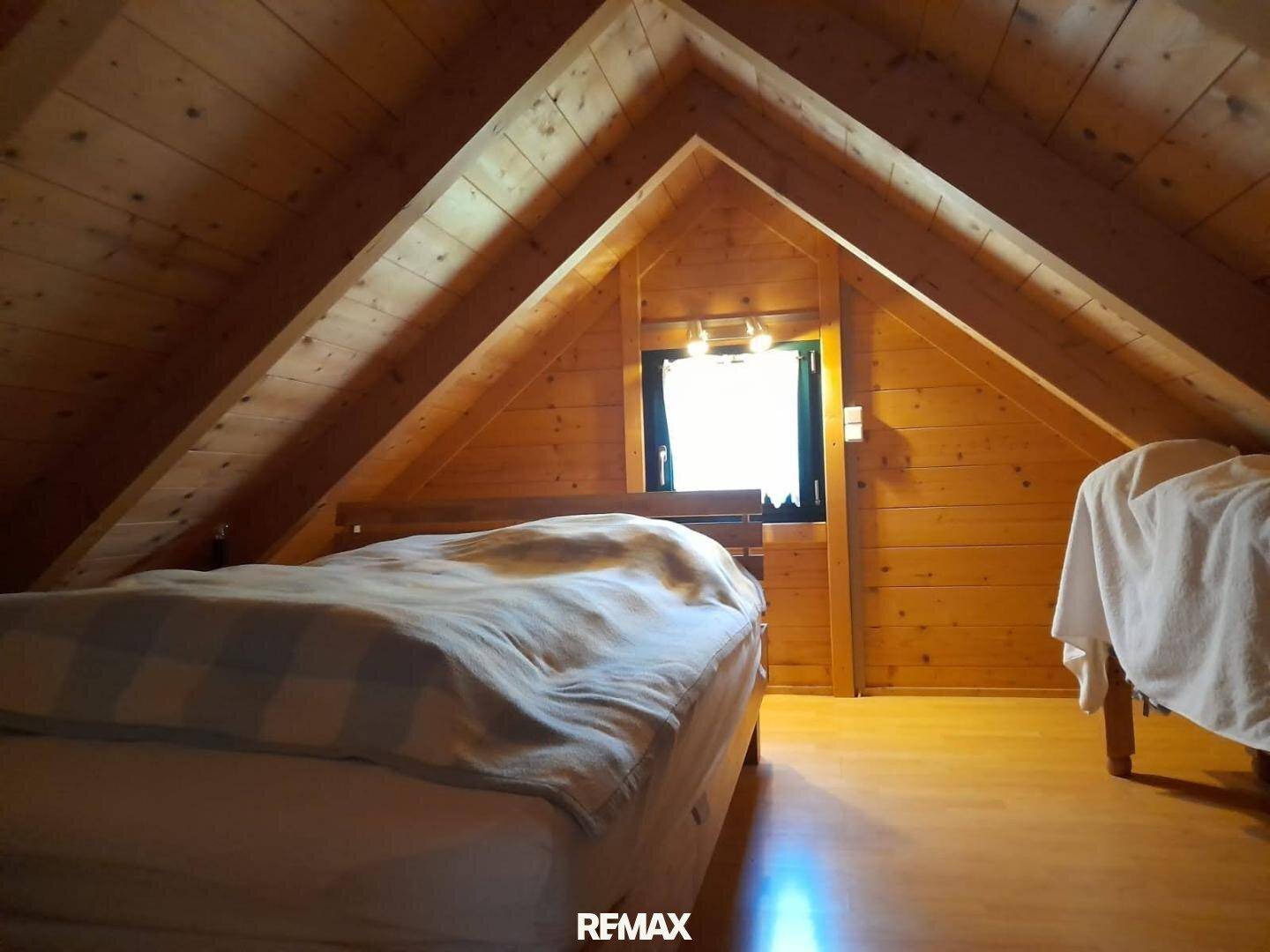 Dachschlafzimmer mit schrägen Holzwänden, sichtbaren Balken und einem Fenster im Giebelbereich.