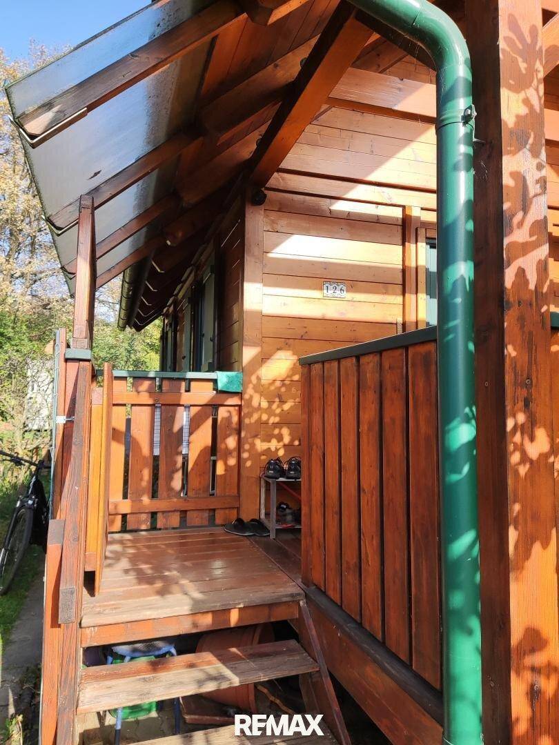 Überdachter Eingangsbereich des Holzhauses mit Holztreppe und grünem Regenrohr im Sonnenlicht.