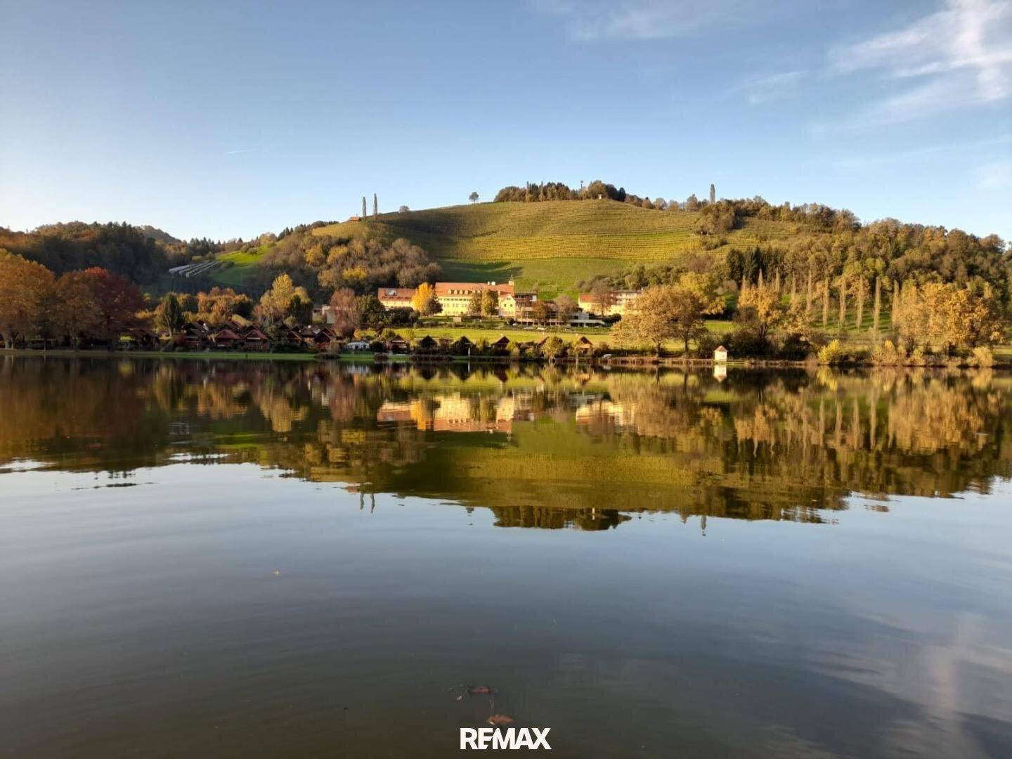 Breiter Panoramablick auf einen ruhigen See mit bewaldeten Ufern und einem Dorf im Hintergrund.