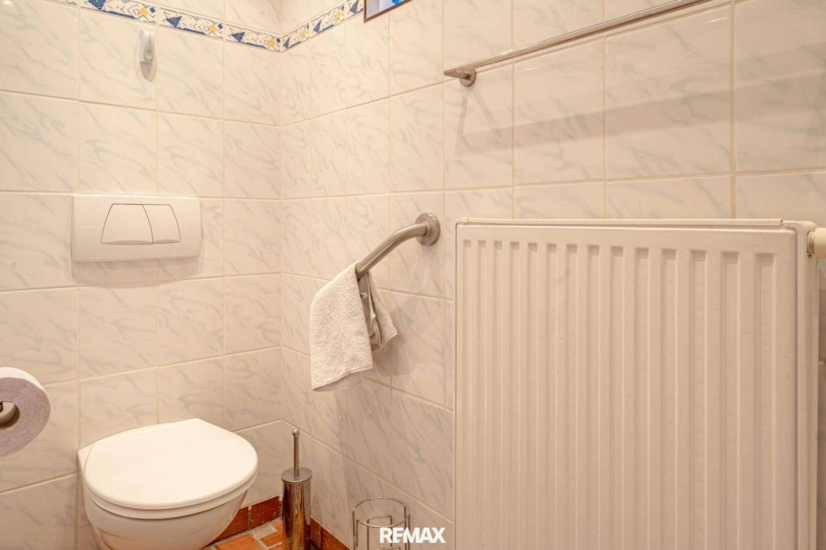 Sauberer Toilettenbereich mit Wand-WC, Heizkörper und Haltegriff, komplett gefliest.