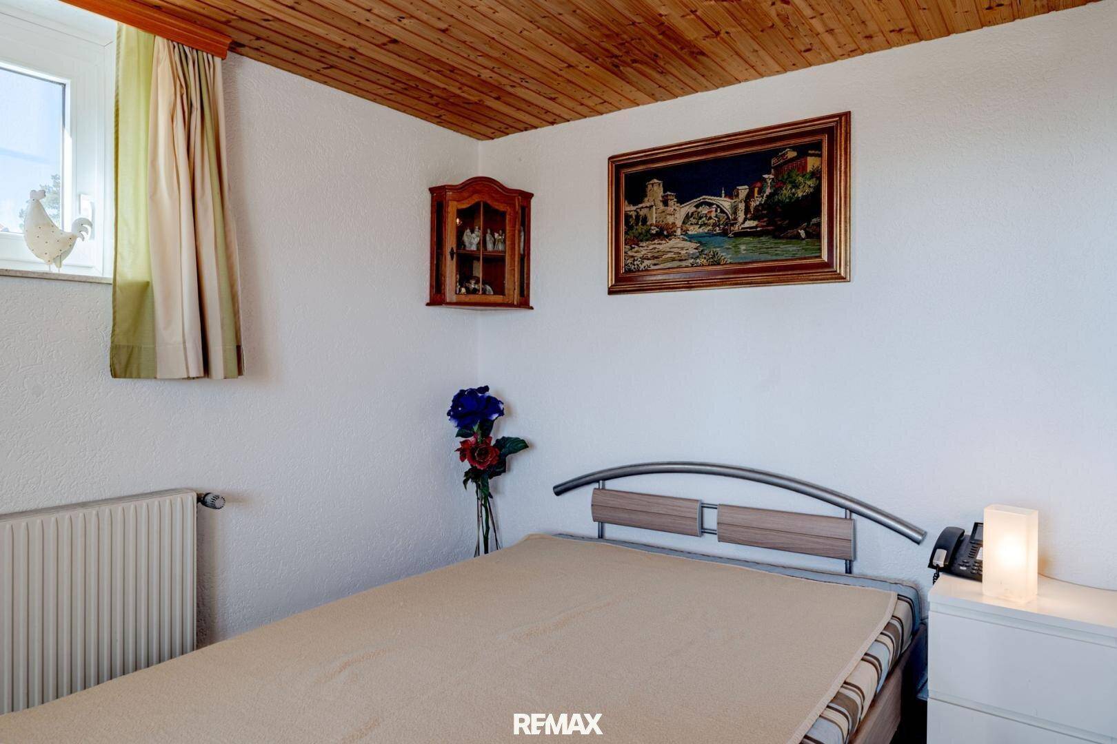 Helles Schlafzimmer mit Holzbalkendecke, Einzelbett und Fenster, das Tageslicht hereinlässt.
