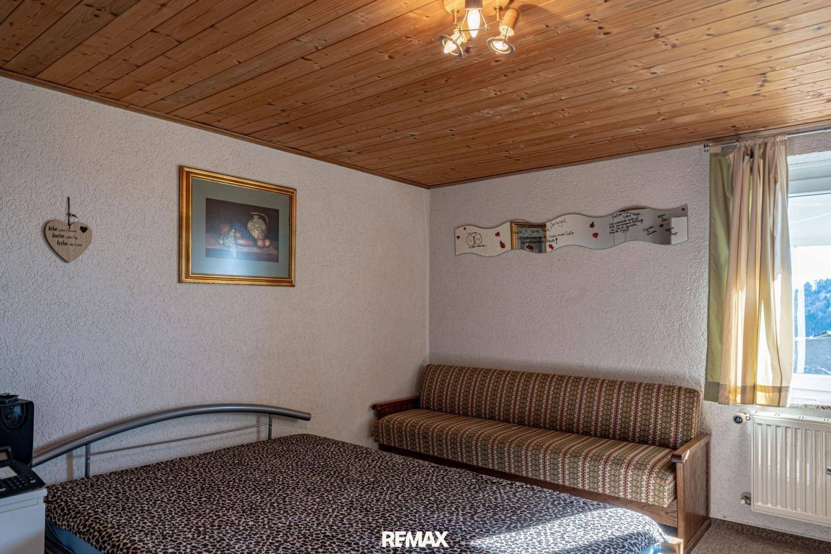 Gemütliches Schlafzimmer mit Holzbalkendecke, Bett und Sofa, hell durch das Fenster.
