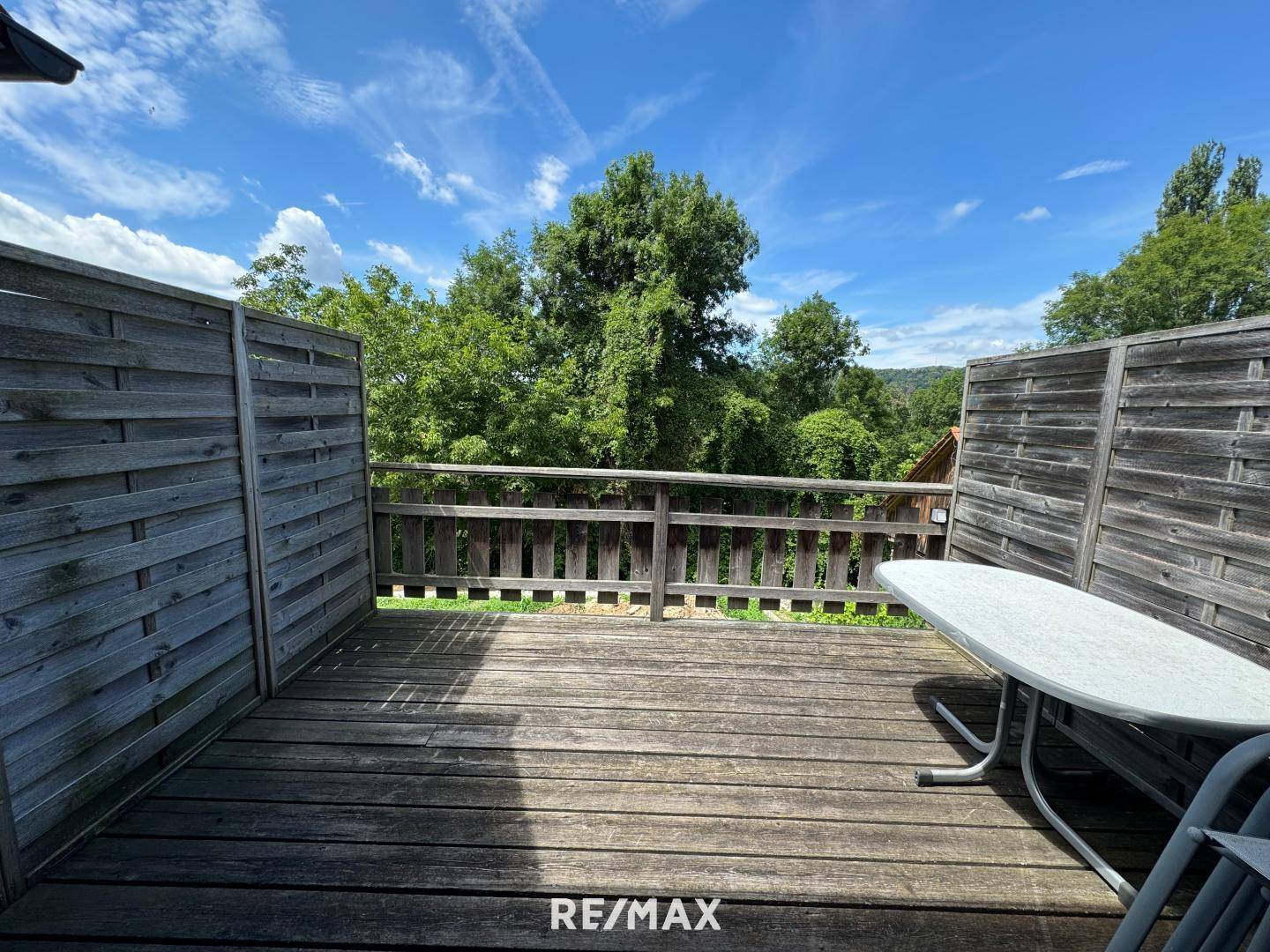 Geräumige Holzterrasse mit Tisch und Stühlen, umgeben von einer Holzwand und üppigem Grün.