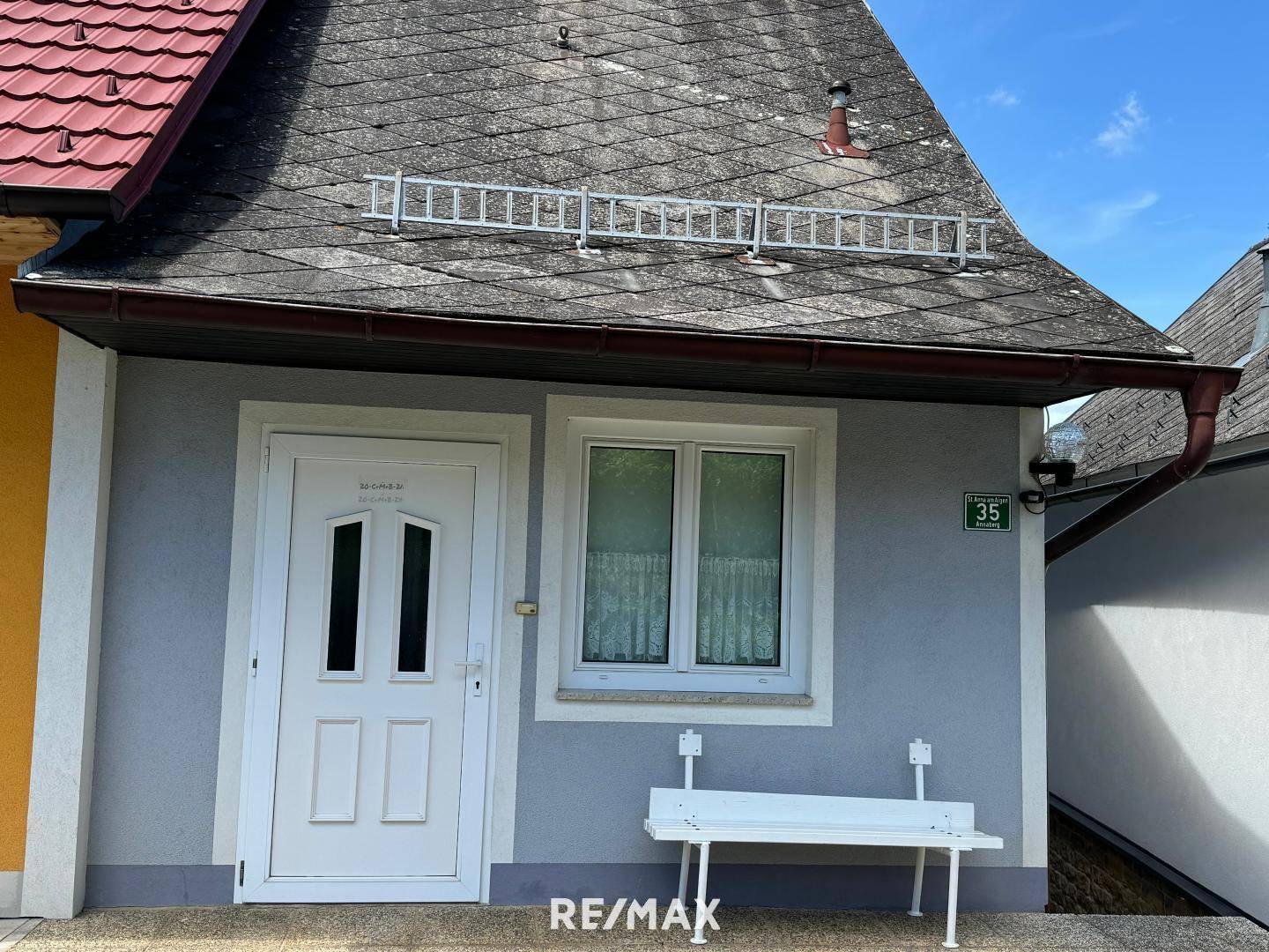 Eingangsbereich eines Hauses mit weißer Tür, Fenster und grauer Fassade unter einem Schindeldach.