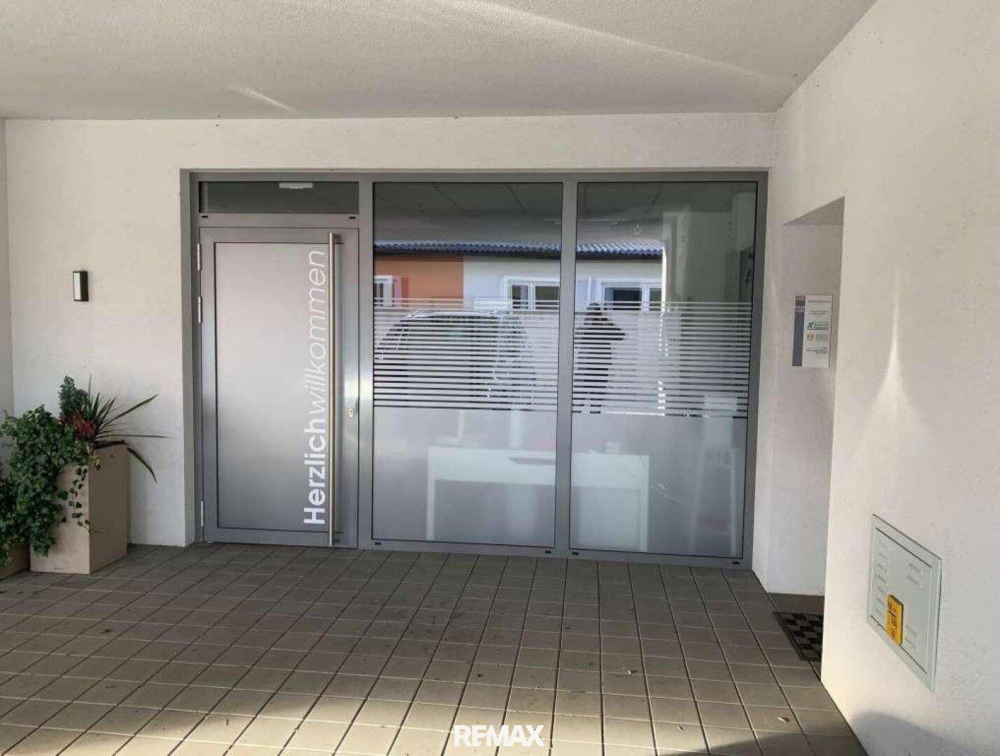 Eingangsbereich mit einer Glastür, die mit "Herzlich willkommen" beschriftet ist, und blickdichten Fenstern.