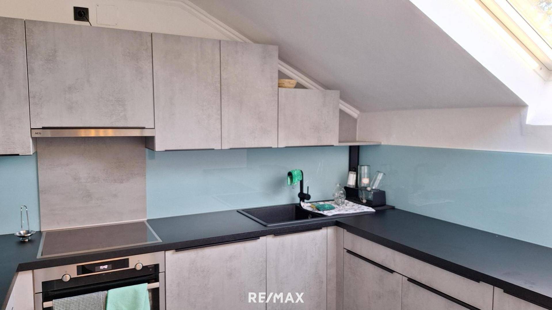 Moderne Küche mit grauen Schränken, dunkler Arbeitsplatte und hellblauer Rückwand im Dachgeschoss.