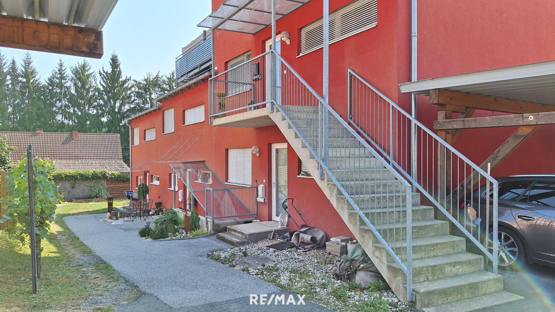 Rote Hausfassade mit Außentreppe, Carport und Blick auf die umliegende Natur.