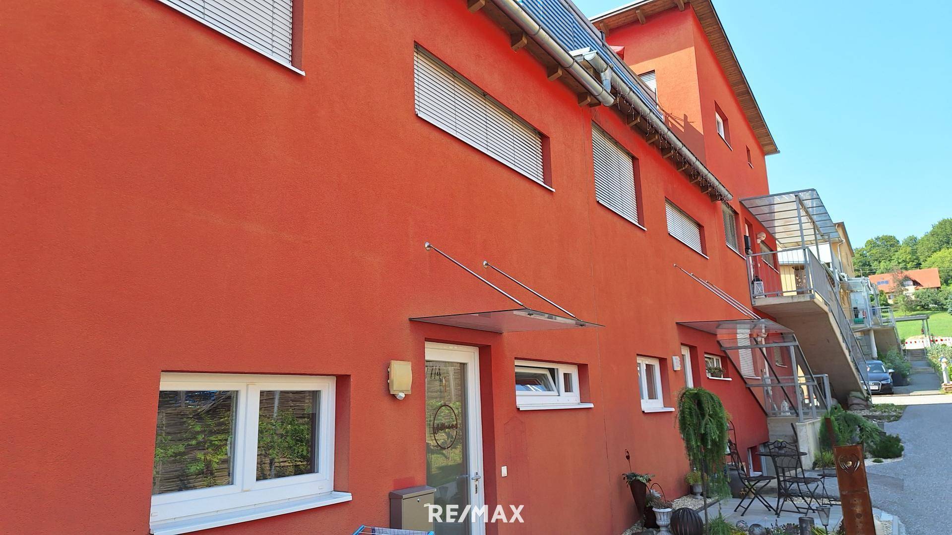 Rote Hausfassade mit Eingangstür, kleinen Fenstern und einem gemütlichen Sitzbereich im Freien.