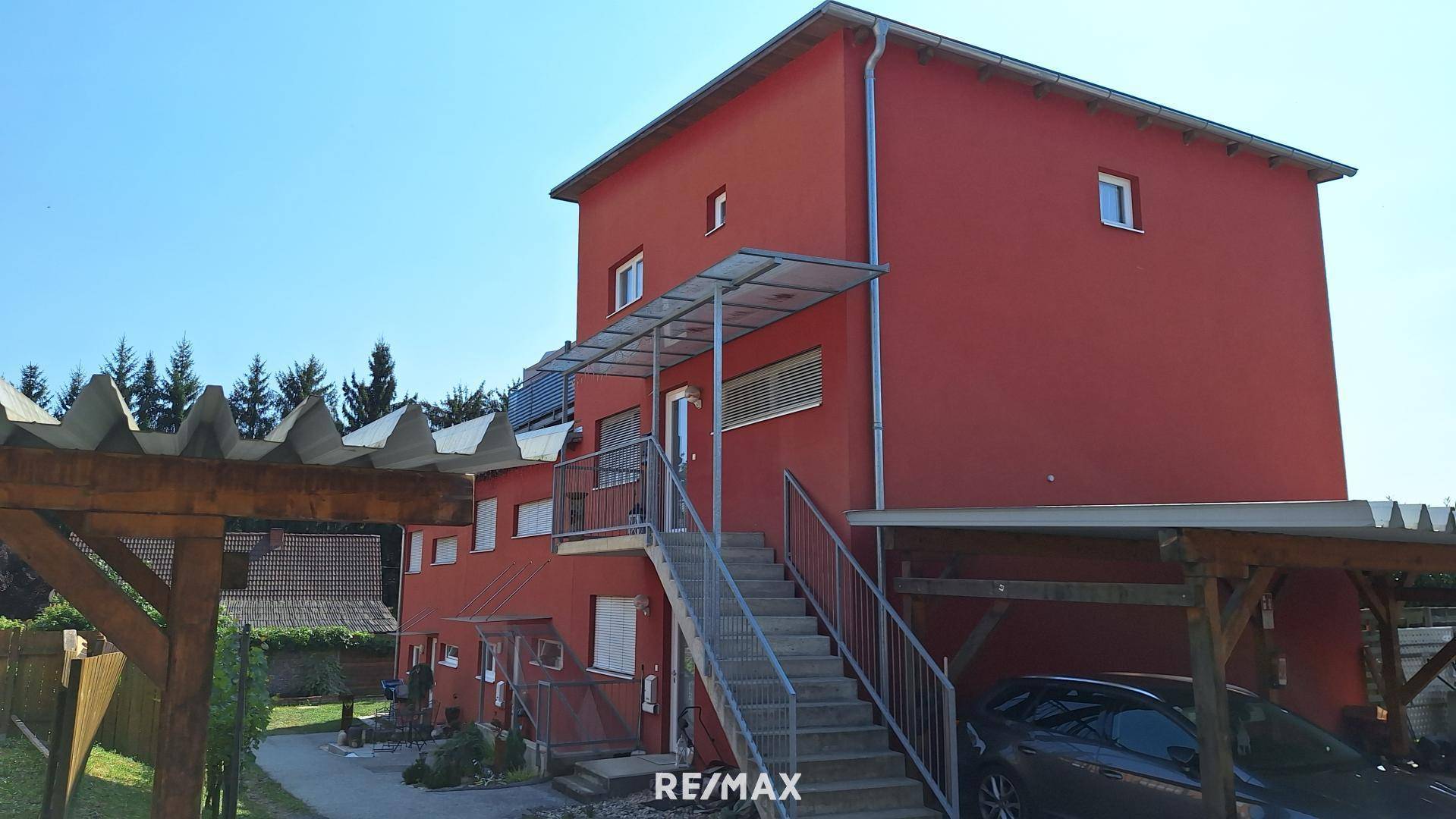 Rotes Mehrfamilienhaus mit Außentreppe, Carport und umgebender grüner Landschaft.