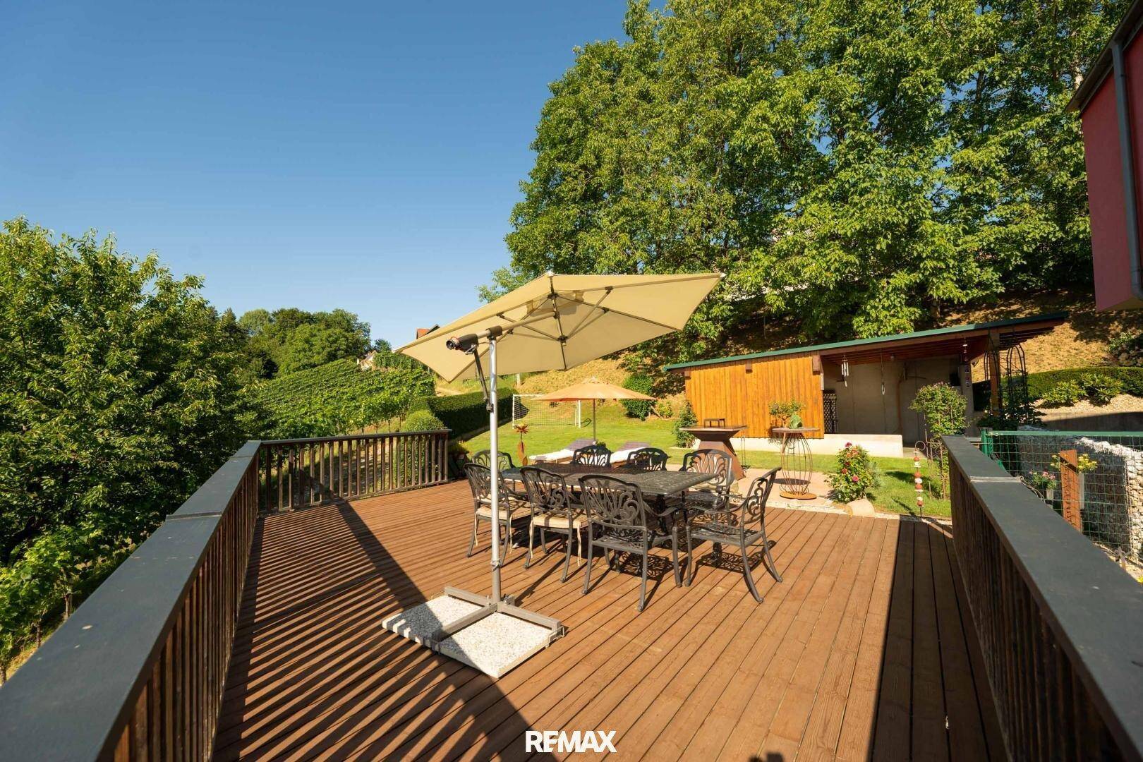 Geräumige Holzterrasse mit Esstisch und Sonnenschirm, umgeben von grüner Natur.