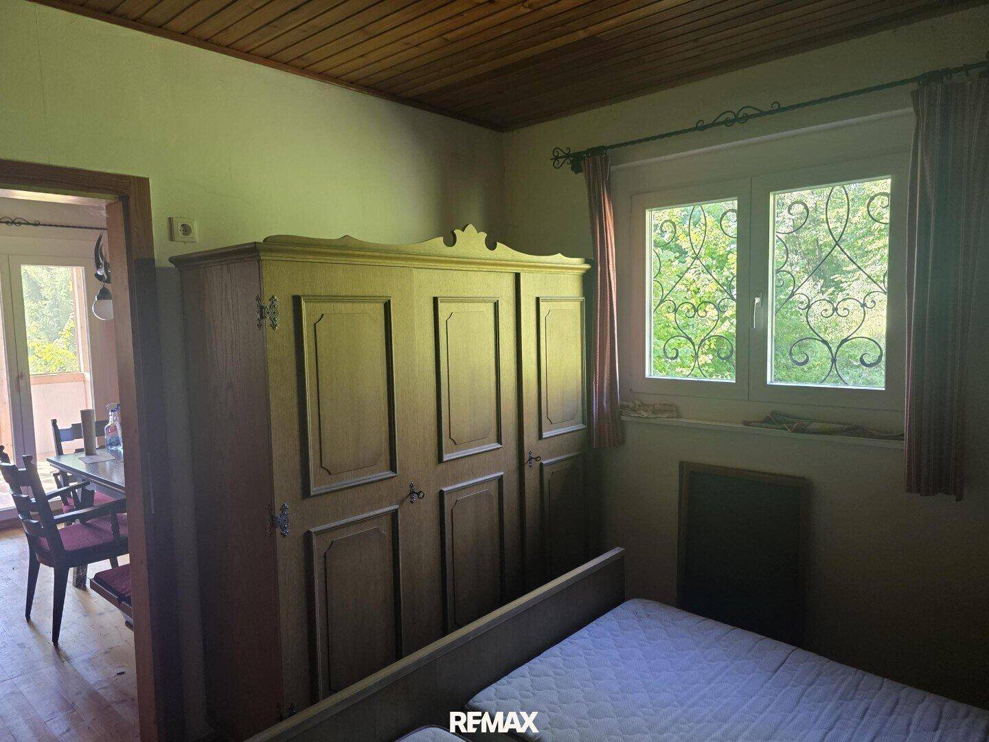 Schlafzimmer mit traditionellem Holzschrank, Holzboden und Fenster mit Blick ins Grüne.