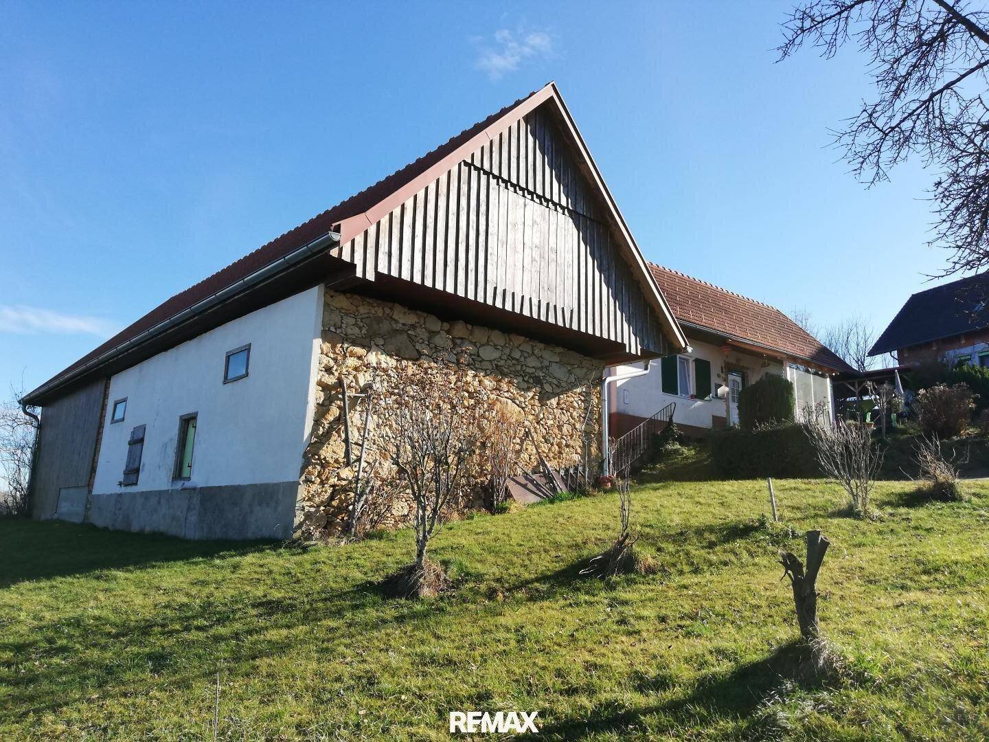 Große Scheune mit Holzwänden und Steinfundament, umgeben von grünen Hügeln unter blauem Himmel.