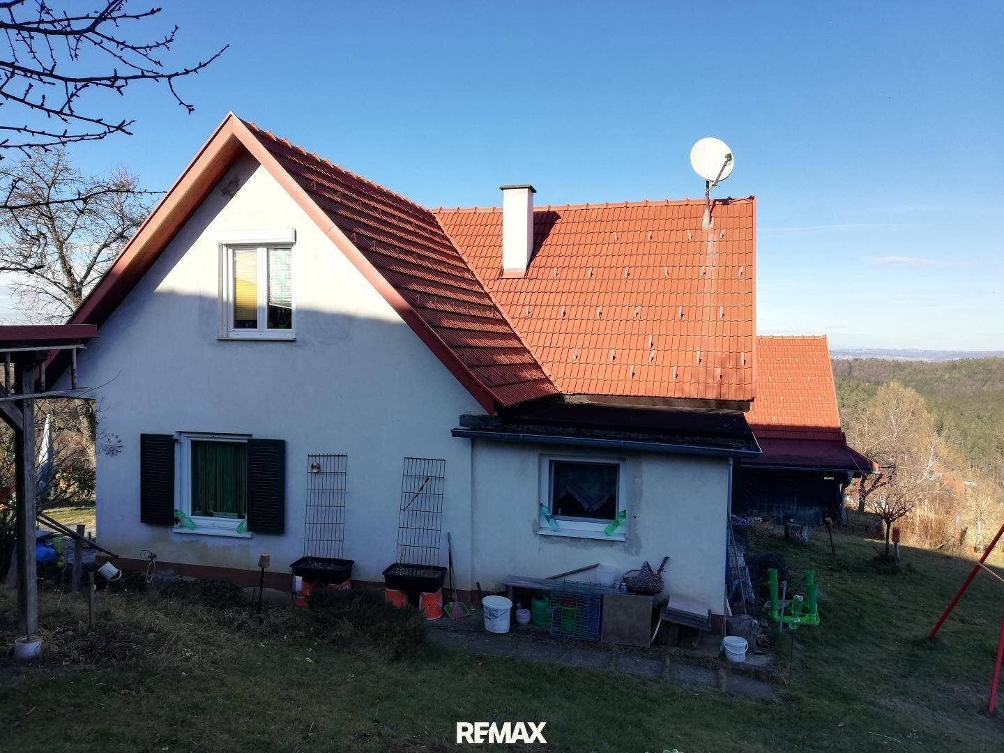 Rückansicht eines ländlichen Hauses mit rotem Ziegeldach und grünen Fensterläden auf einem Hügel.