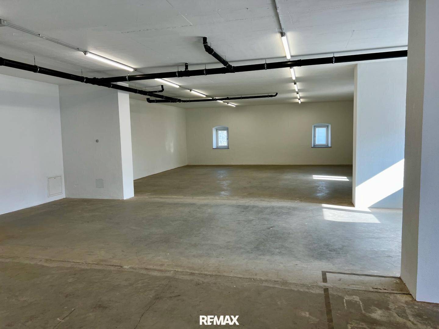 Großer, heller Gewerberaum mit mehreren kleinen Fenstern, Betonboden und sichtbaren Deckeninstallationen.