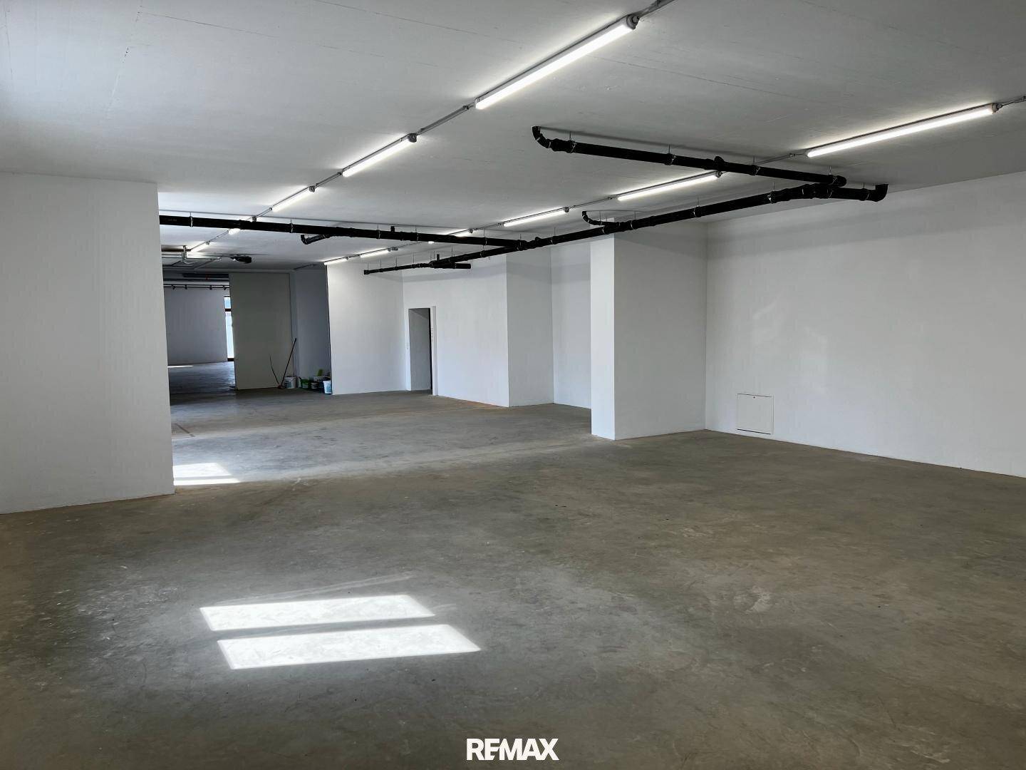 Geräumiger, leerer Gewerberaum mit Betonboden, weißen Wänden und sichtbaren Leitungen an der Decke.