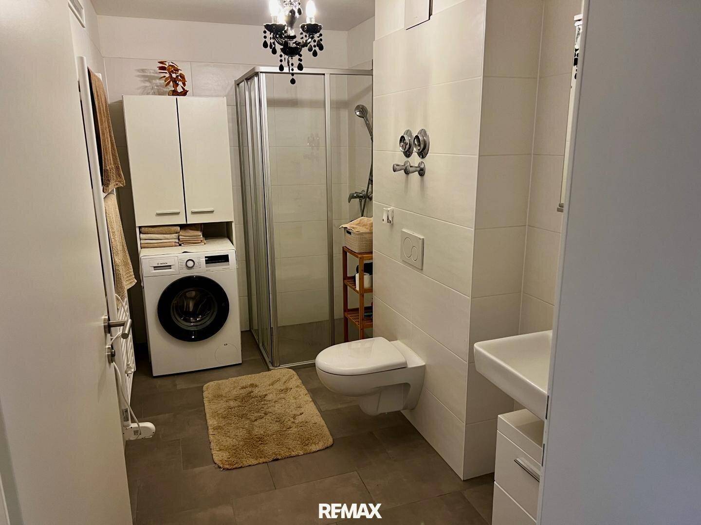 Modernes Badezimmer mit Dusche, WC, Waschbecken und integrierter Waschmaschine, hell gefliest.