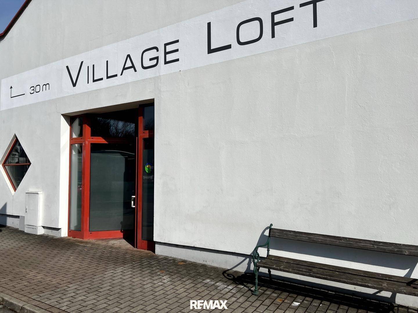 Eingangsbereich des Gebäudes 'Village Loft' mit roter Tür und einer Holzbank davor an einem hellen Tag.