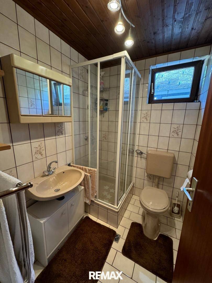 Gefliestes Badezimmer mit Dusche, Waschbecken und WC, im Stil der 80er Jahre.
