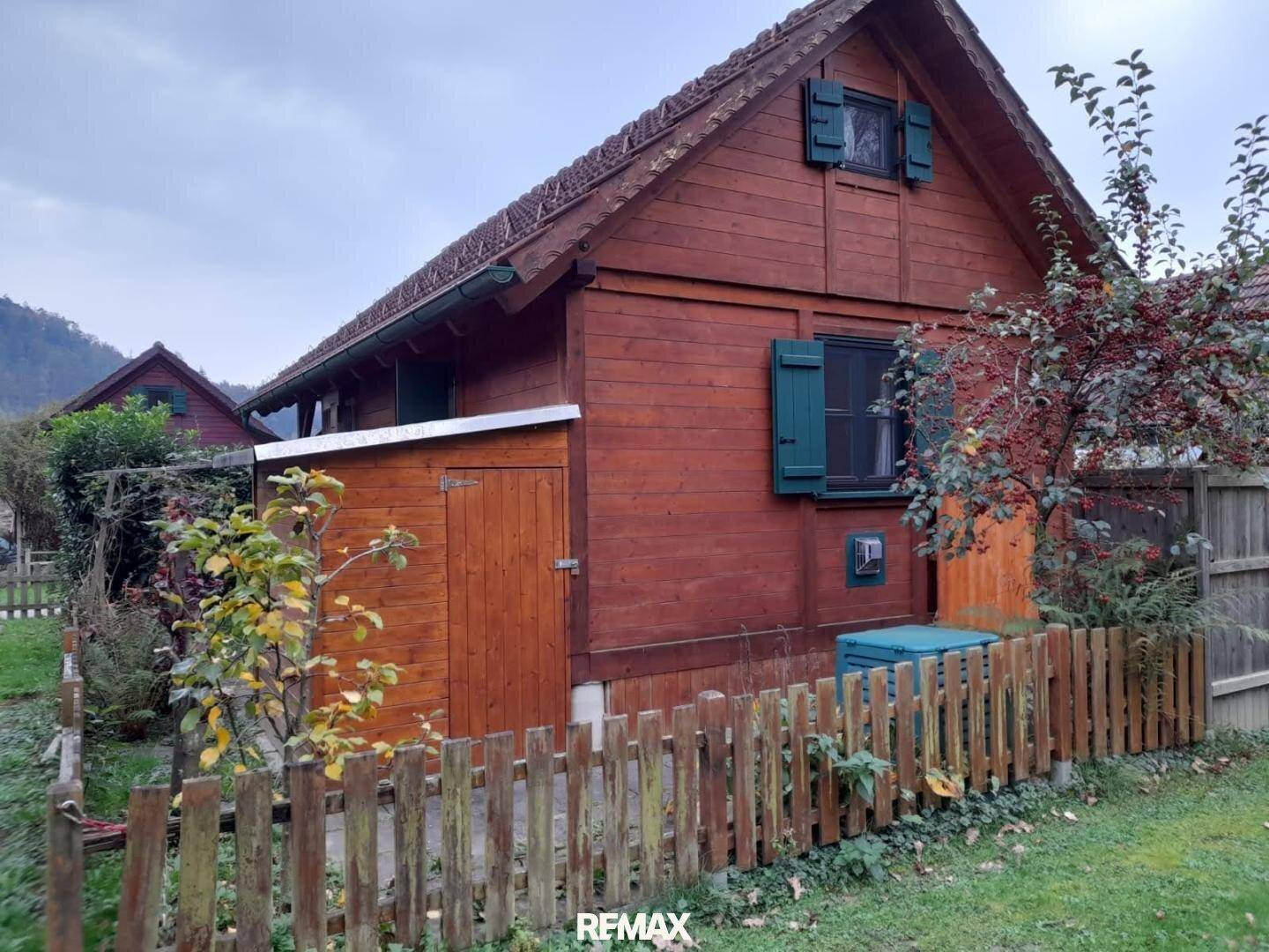 Rückseite eines Holzhauses mit grünem Fensterladen und kleinem Schuppen in ländlicher Umgebung.