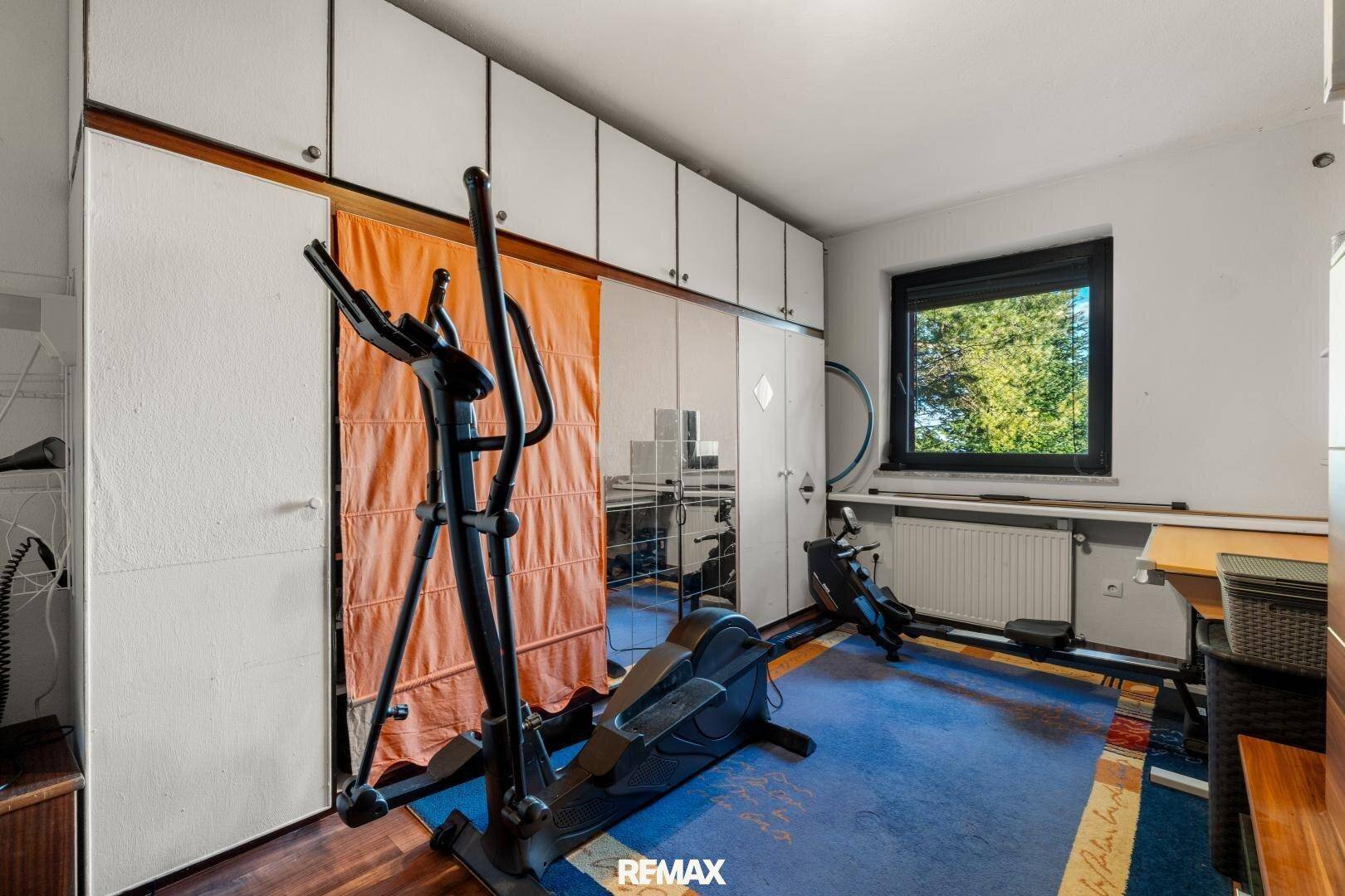 Fitnessraum mit Trainingsgeräten, Teppichboden und Einbauschränken entlang einer Wand.