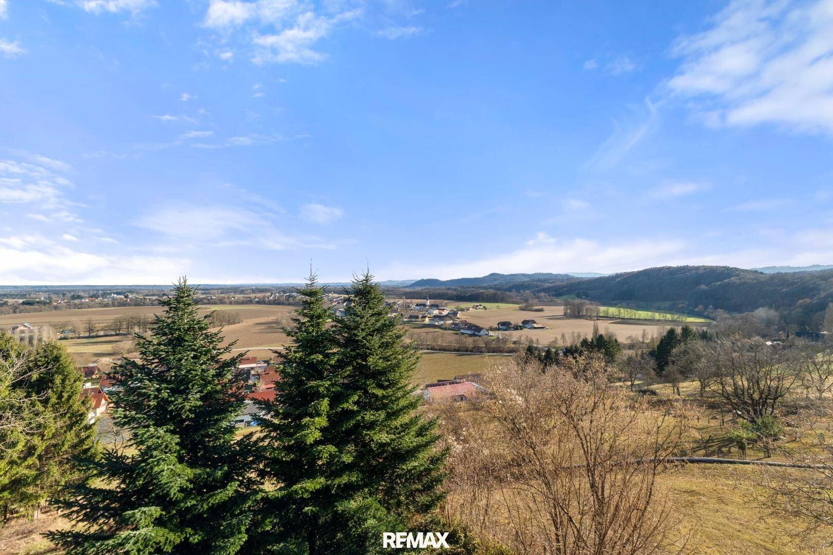 Weitläufiger Blick über die hügelige Landschaft mit Bäumen und vereinzelten Häusern unter blauem Himmel.