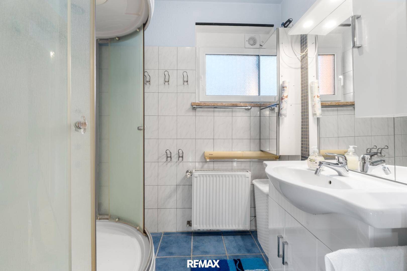 Badezimmer mit Duschkabine, Waschbecken und Fenster, gefliest in Weiß und Blau.
