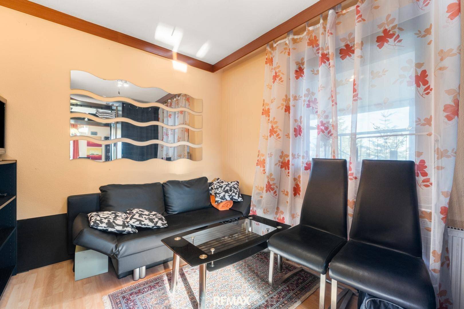 Gemütliches Wohnzimmer mit schwarzem Sofa, Glastisch und floralen Vorhängen, hell durch ein Fenster.