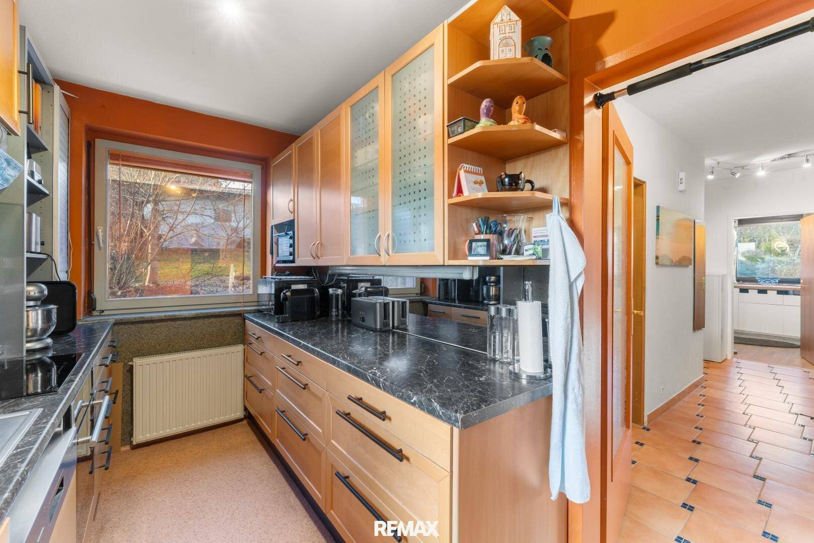 Küche mit Holzschränken, dunkler Arbeitsplatte und Fenster, das Licht hereinlässt, mit Fliesenboden im Flur.