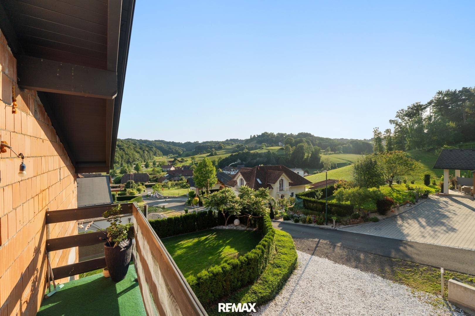 Blick vom Balkon auf die umliegende grüne Landschaft und benachbarte Häuser unter blauem Himmel.