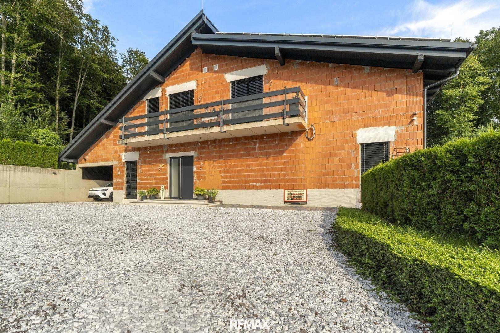 Neubauhaus mit unverputzten Ziegelwänden und Balkon im ländlichen Umfeld, umgeben von Bäumen.