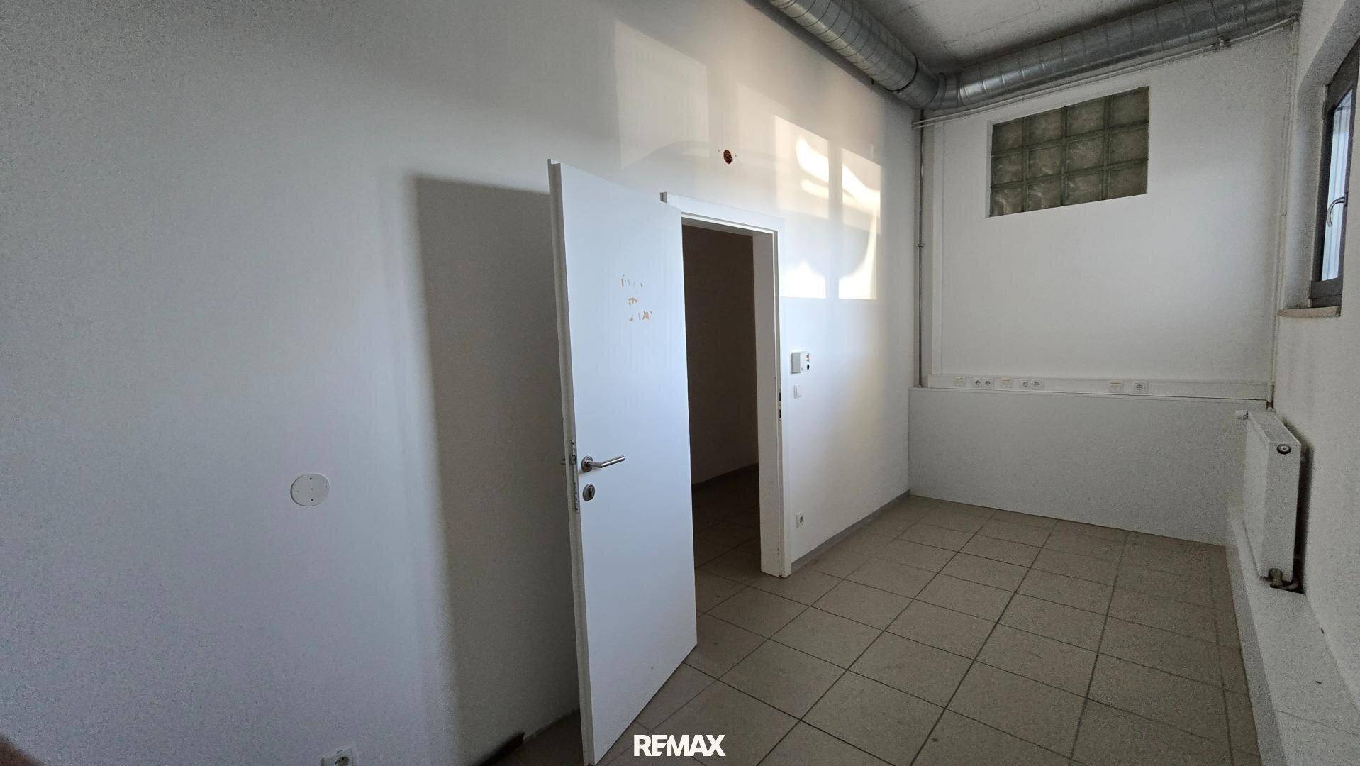Kleiner Büroraum mit weißer Tür, Fliesenboden und einem Fenster mit Glasbausteinen für Tageslicht.