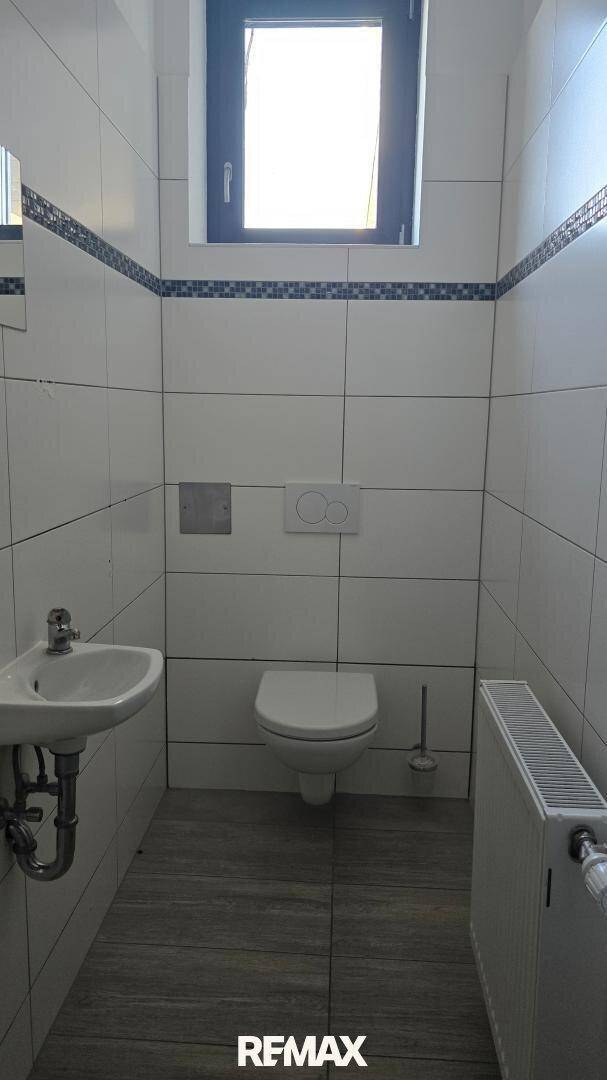 Helles Gäste-WC mit Fenster, wandhängender Toilette und Waschbecken, ausgestattet mit weißen Fliesen.